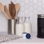 Keurig Milk frother on a countertop with bottles of cold milk and kitchen tools against a hexangal white tile backsplash.