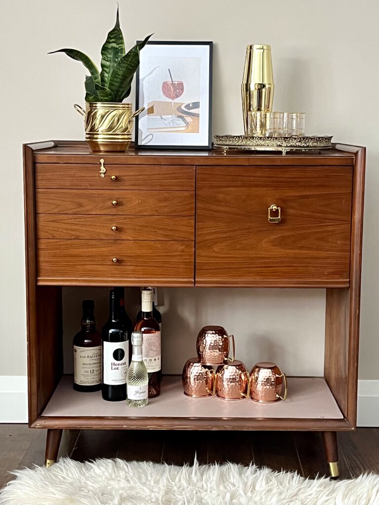 A teak mid century bar stand accessorized with copper cups, metal cocktail shaker, and art makes for sustainable style.
