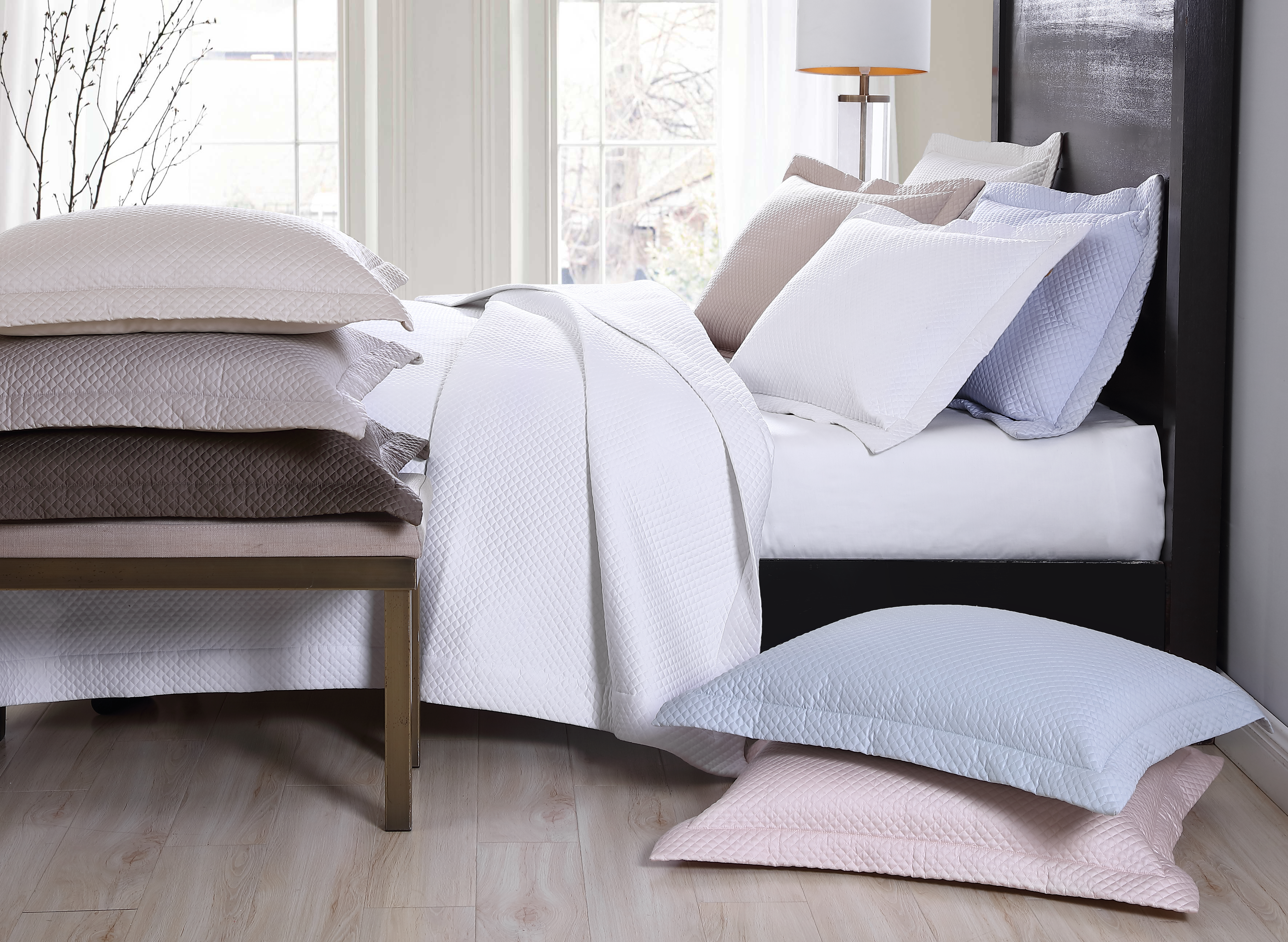 sunshine from a window spills onto a modern bed frame made of wood made with fresh crisp white bedding and several pillows in different shades of blue, beige, pink and white