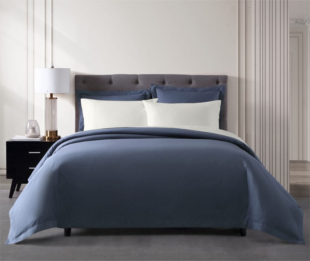 A queen size bed against a coffered cream wall dressed in navy blue linen bedding and piled high with crisp, fluffy white pillows.