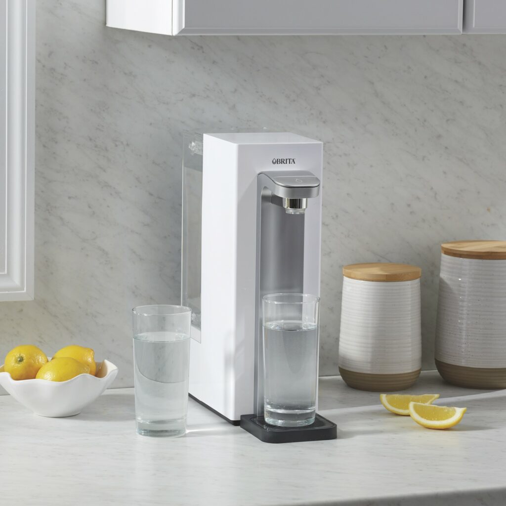 A slim line Brita Hub Compact water filter and dispenser on a counter with a fresh glass of water and sliced lemons beside it as an example of a simple way to reduce plastic use.